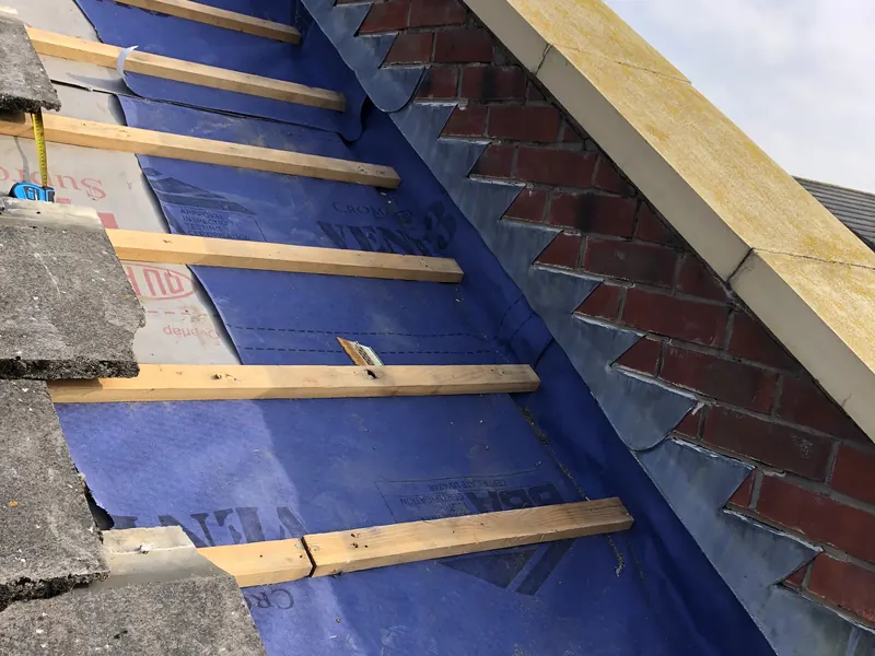 Stepped lead flashing against a brick parapet wall with coping stones, above new blue roofing membrane and timber battens
